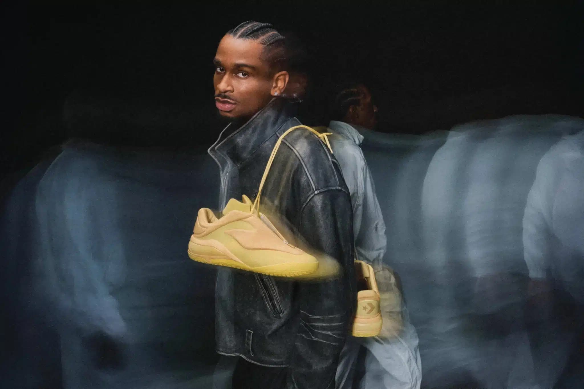 A person wearing a black jacket holds a single tan sneaker over their shoulder against a dark, blurred background.