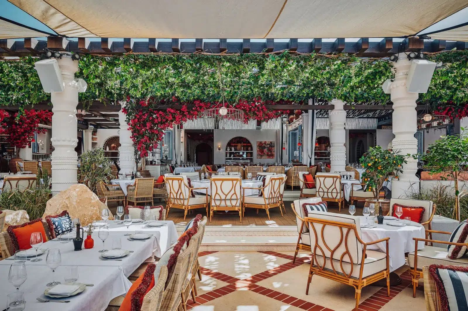 Outdoor restaurant with white tablecloths, wicker chairs, and leafy green and red floral decor under a shaded canopy.
