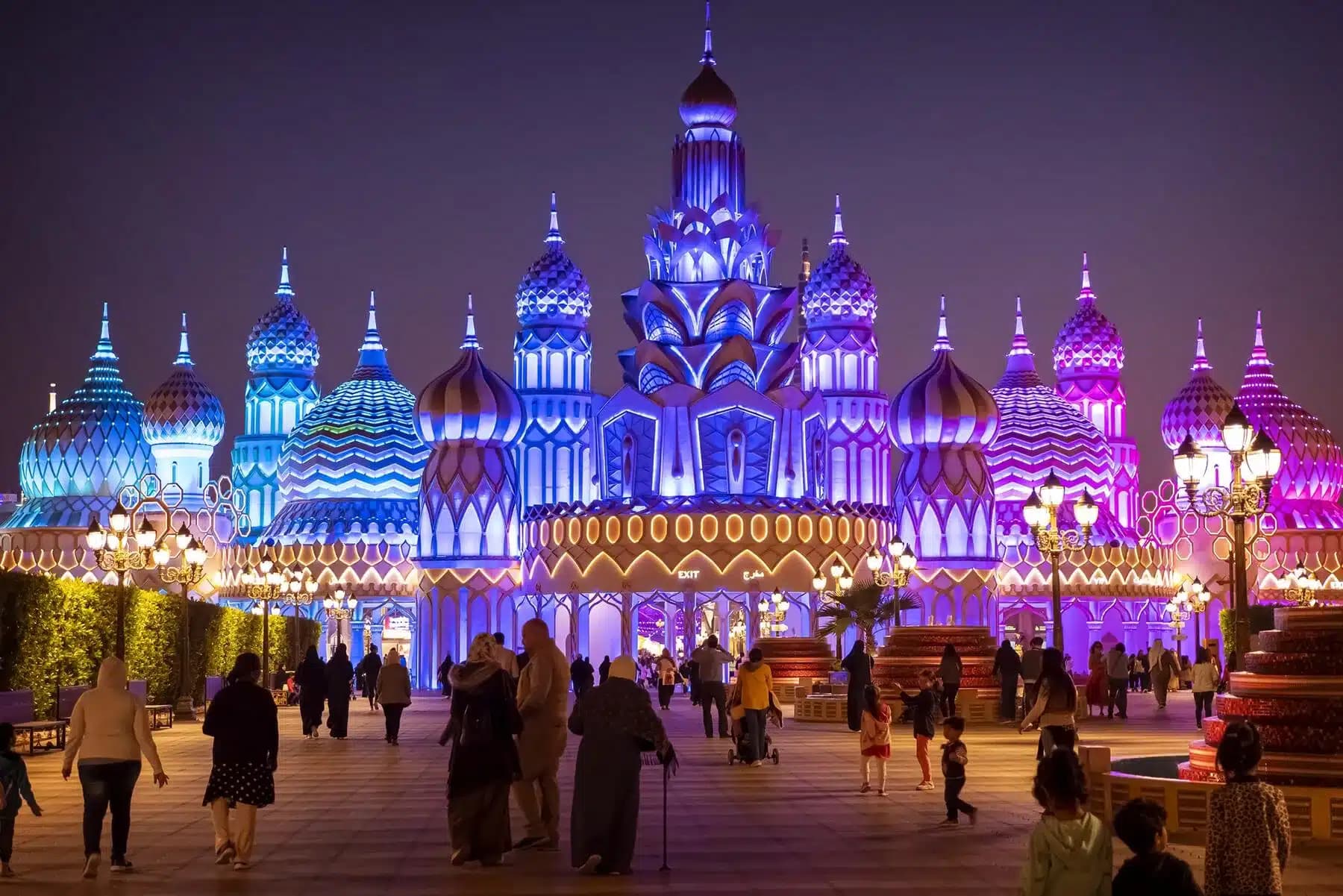 An ornate, brightly illuminated building with domes and spires at night, surrounded by walking visitors.