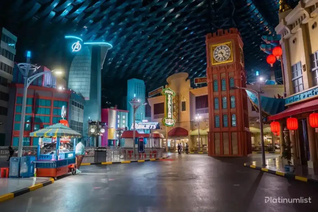 A lively indoor theme park scene with colorful buildings, amusement signs, and a food cart on a wide pathway.