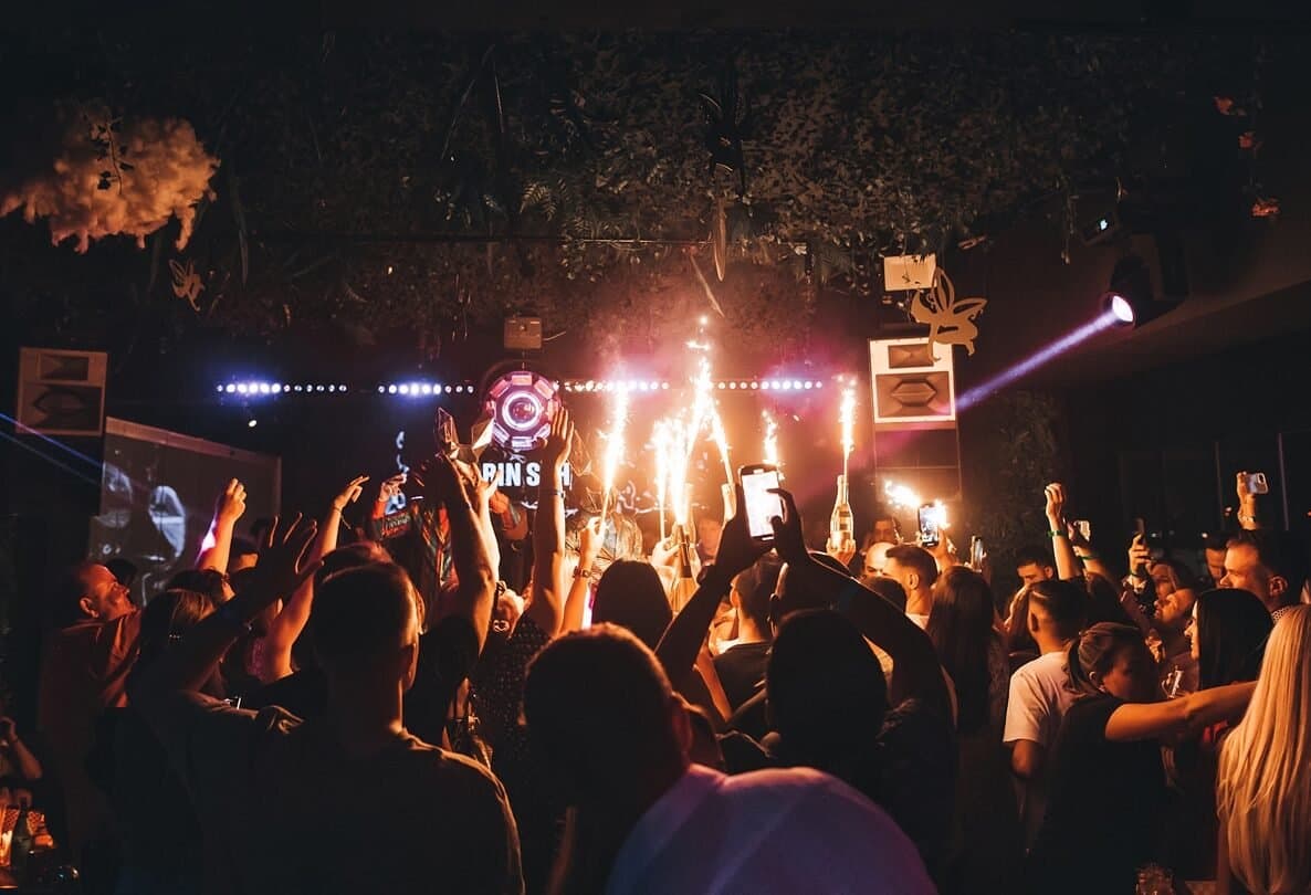 A lively crowd at a nightclub, holding up phones and drinks as sparklers light up the room.