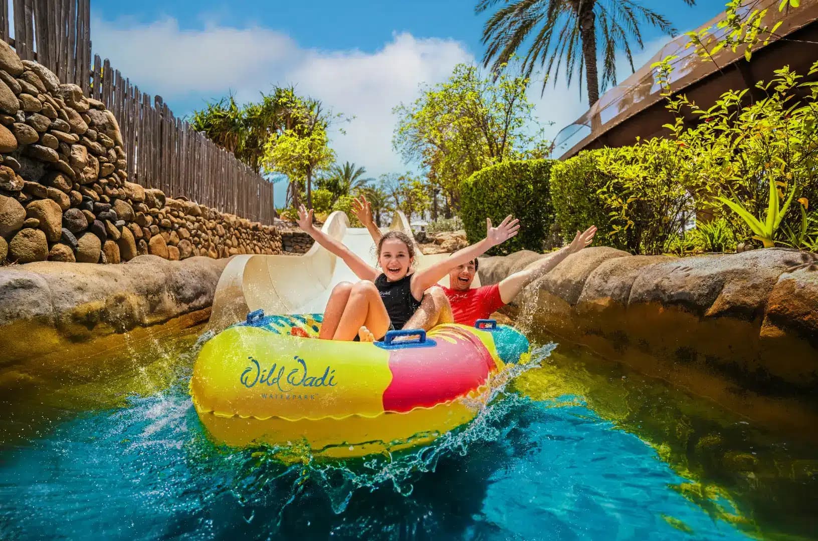 Two people joyfully riding a colorful inflatable raft on a water slide at a tropical water park.