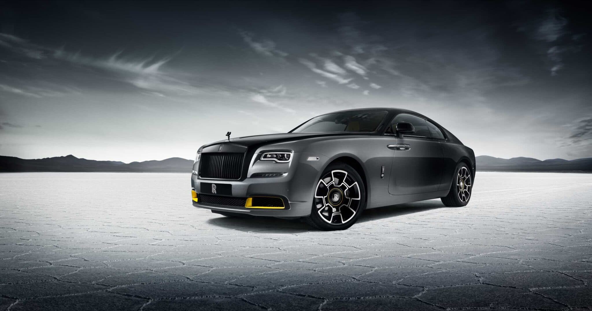 A sleek, dark gray luxury car with distinctive design and intricate wheels, set against a vast, barren landscape.