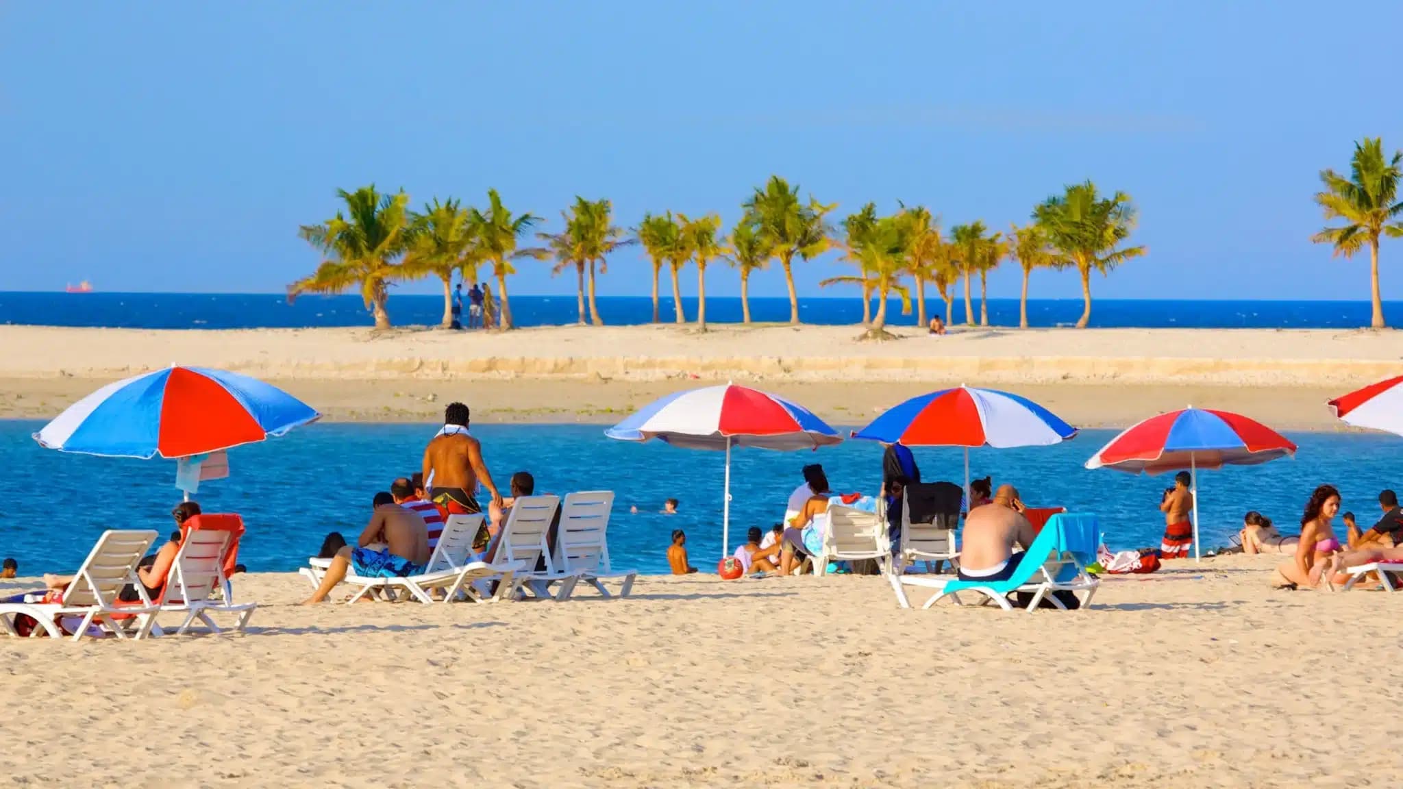 People relax on sun loungers under red and white umbrellas on a sandy beach, with palm trees and the sea in the background.