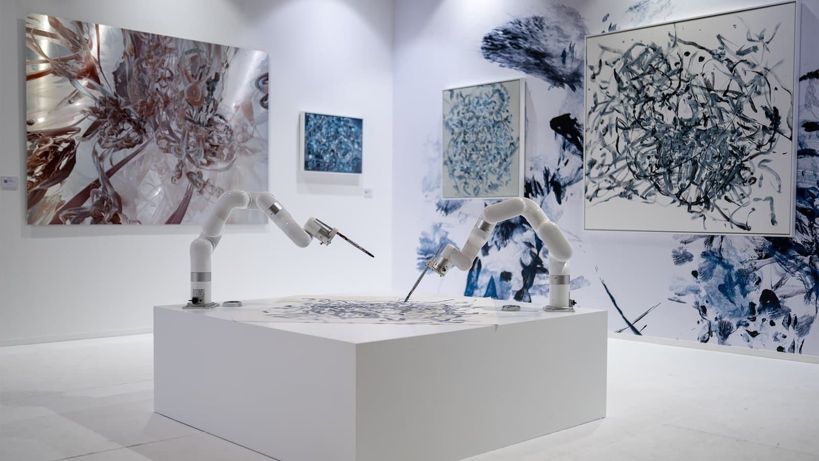 Robotic arms create abstract art on a tabletop in a gallery with abstract paintings on the walls.