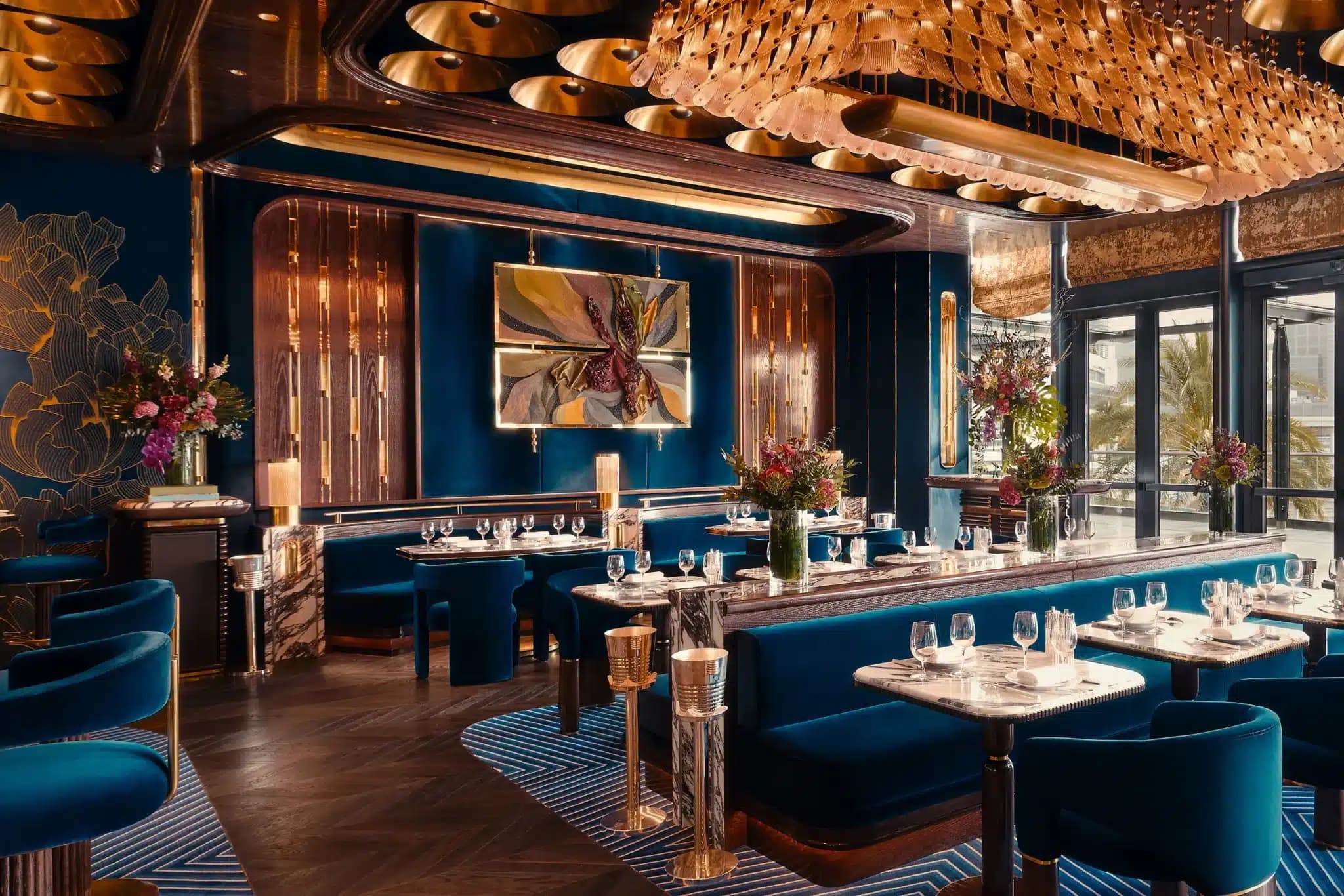 Luxurious restaurant interior with blue velvet seating, marble tables, ornate lighting, and floral arrangements.