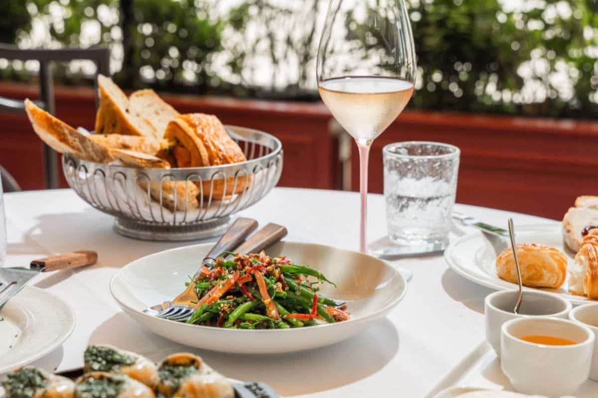 Table with a dish of vegetables and chopsticks, a basket of bread, a glass of wine, and various side dishes.