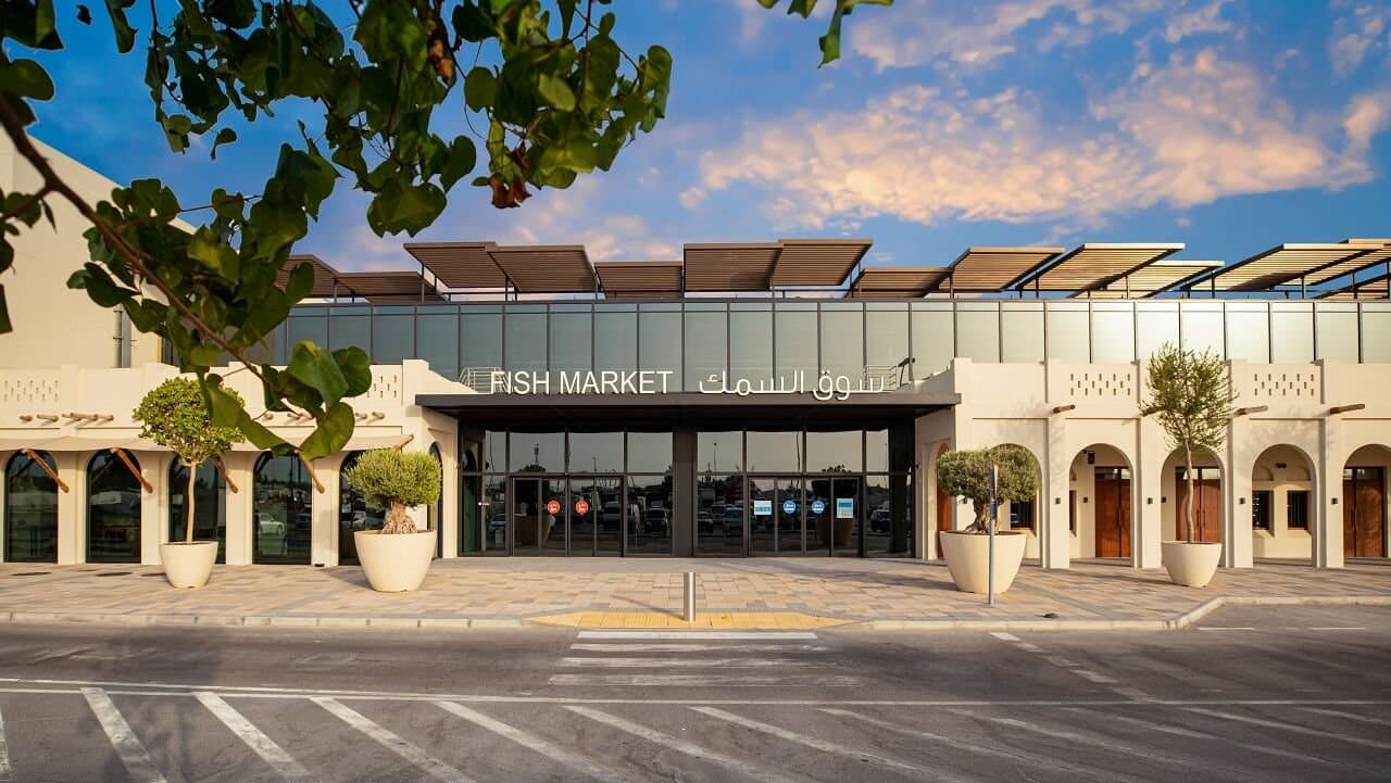 Modern building labeled "Fish Market" with large windows, surrounded by trees and potted plants, under a partly cloudy sky.