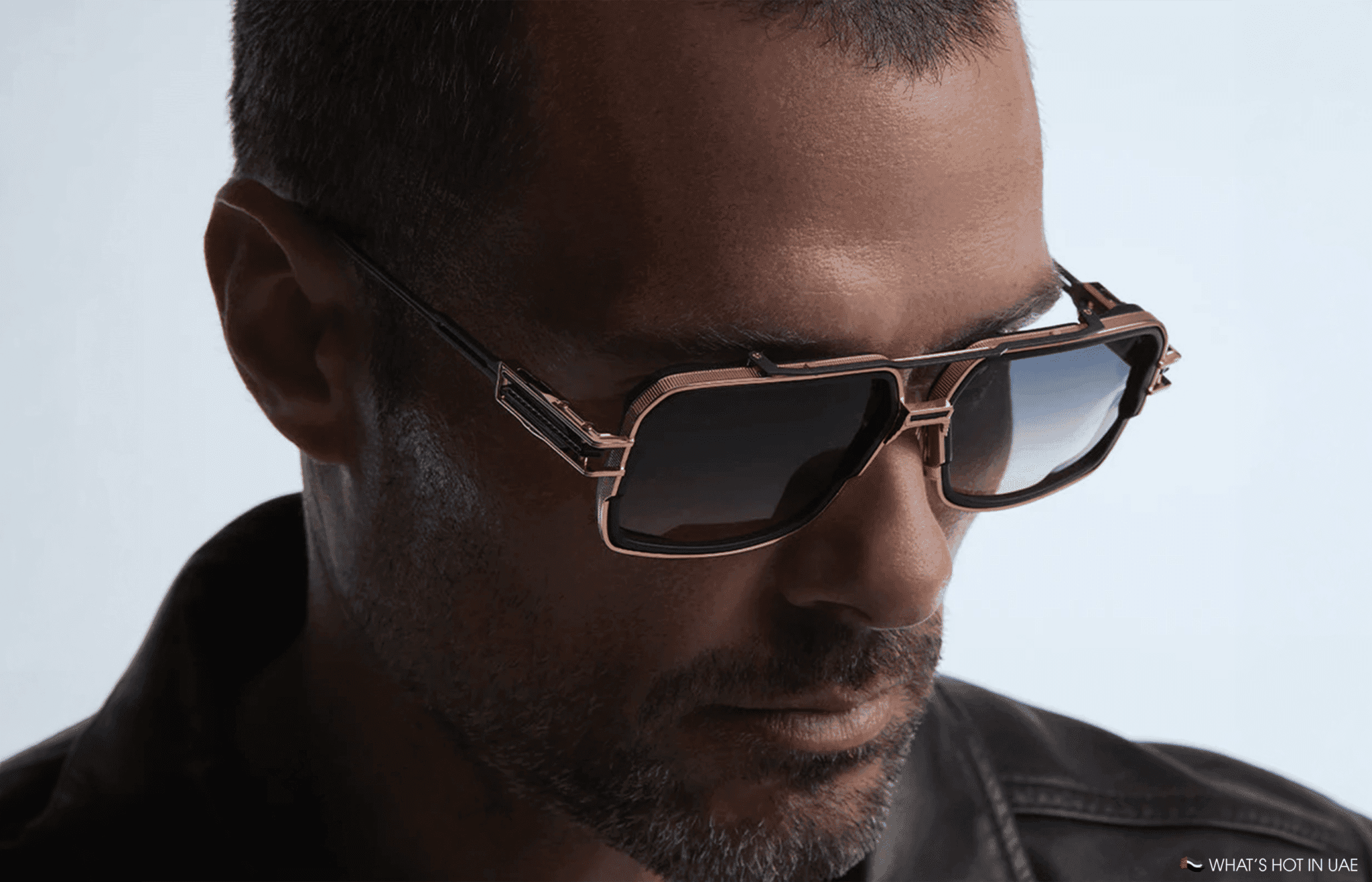 Close-up of a man wearing gold-framed sunglasses and a dark jacket against a light background