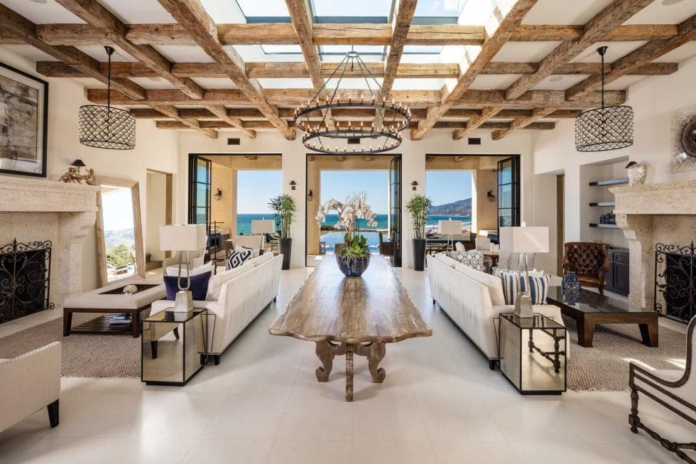 Luxurious living room with wooden ceiling beams, a long wooden table, sofas, chandeliers, and large windows revealing a scenic view.
