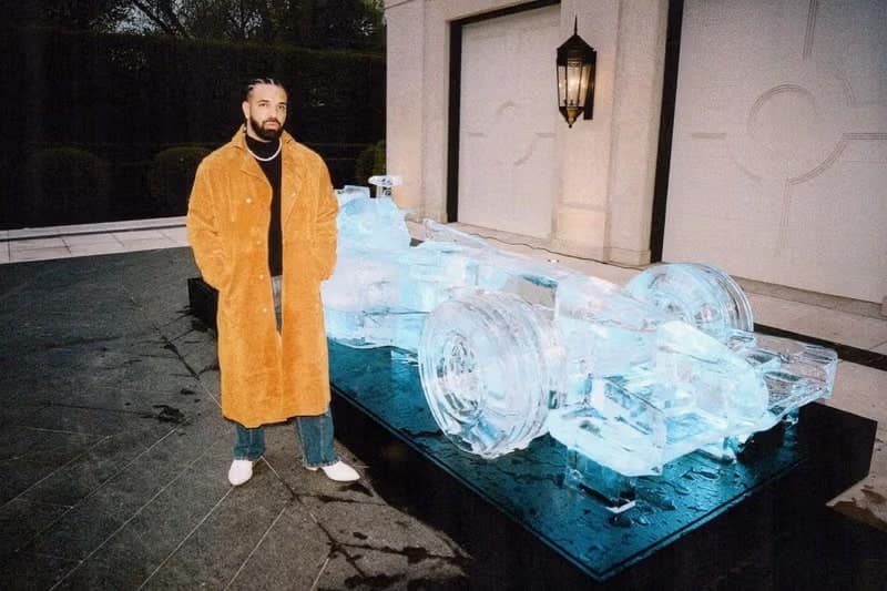 A man in a tan coat stands beside a large ice sculpture of a car outdoors at night