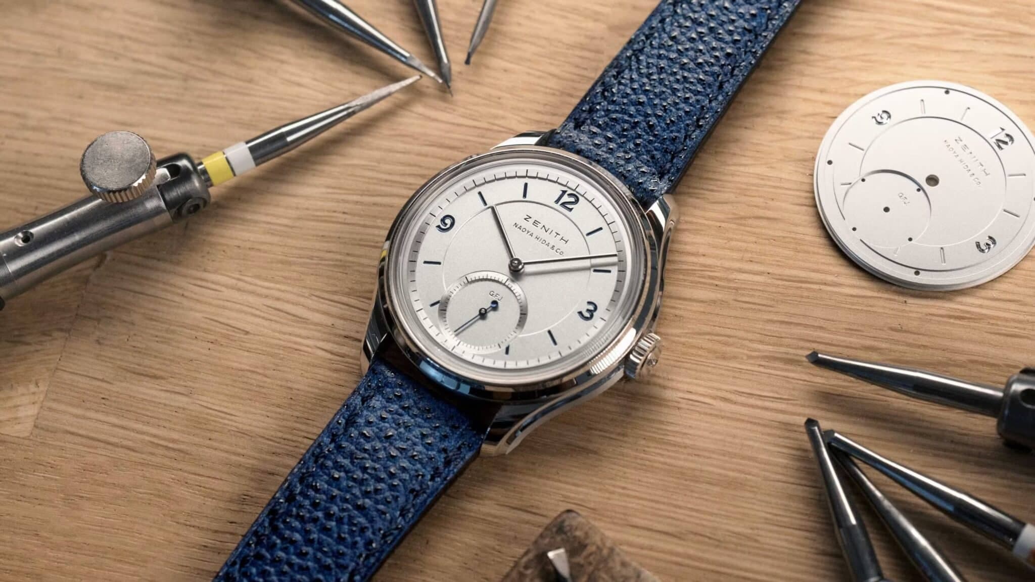 Open watch with blue strap on a wooden workbench, surrounded by watchmaker tools and a separate dial