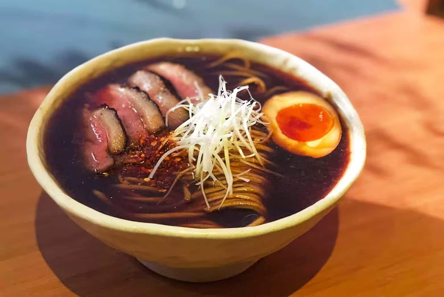 Bowl of dark broth ramen topped with sliced meat, soft-boiled egg, and shredded scallions on a wooden table
