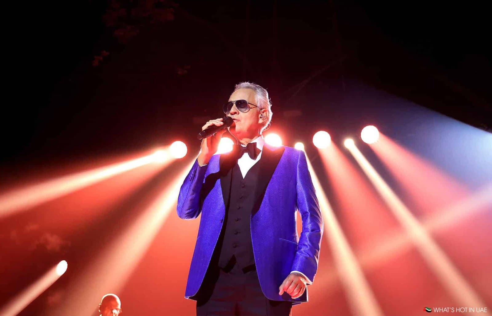 An older man in a blue suit and sunglasses sings into a microphone under bright stage lights