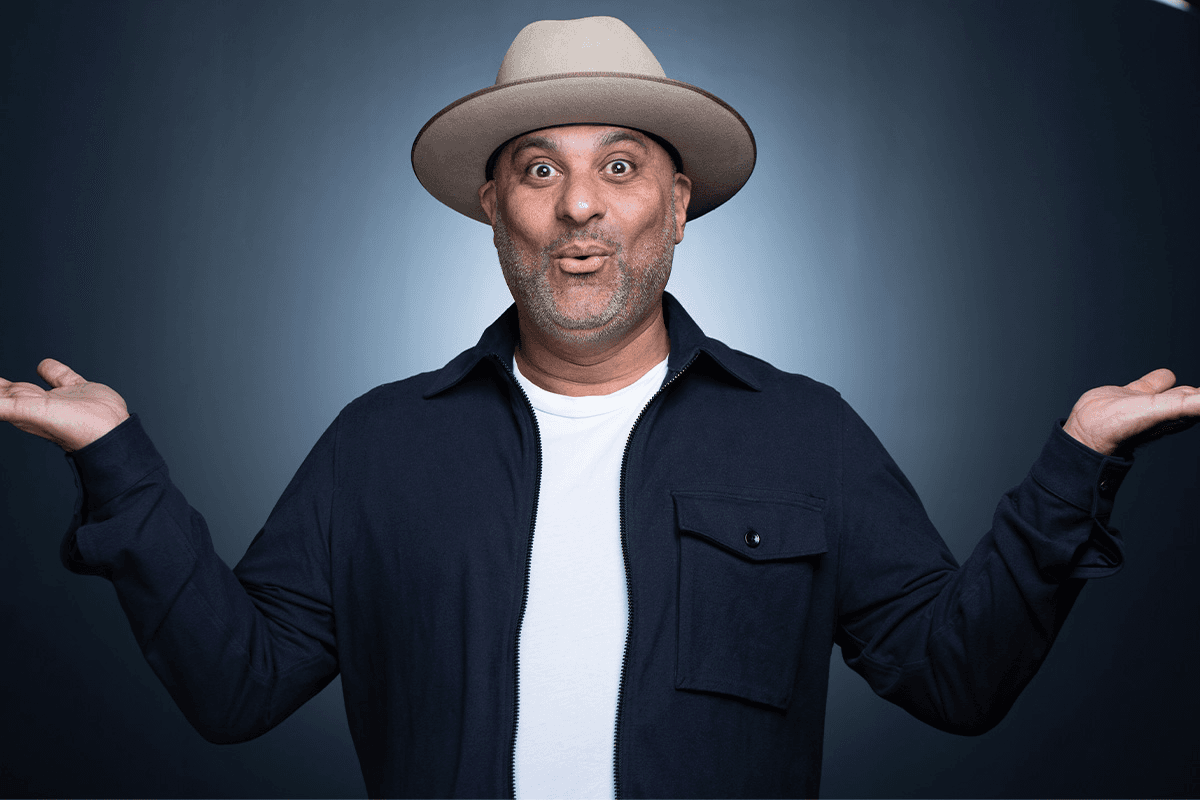Smiling man in a beige hat and dark jacket shrugs with both hands raised against a blue background
