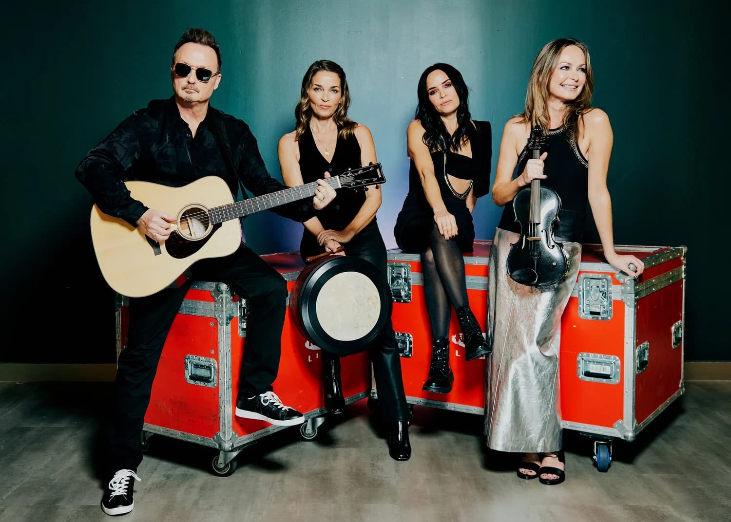 Four musicians pose with guitar and violin on red road cases against a dark teal wall