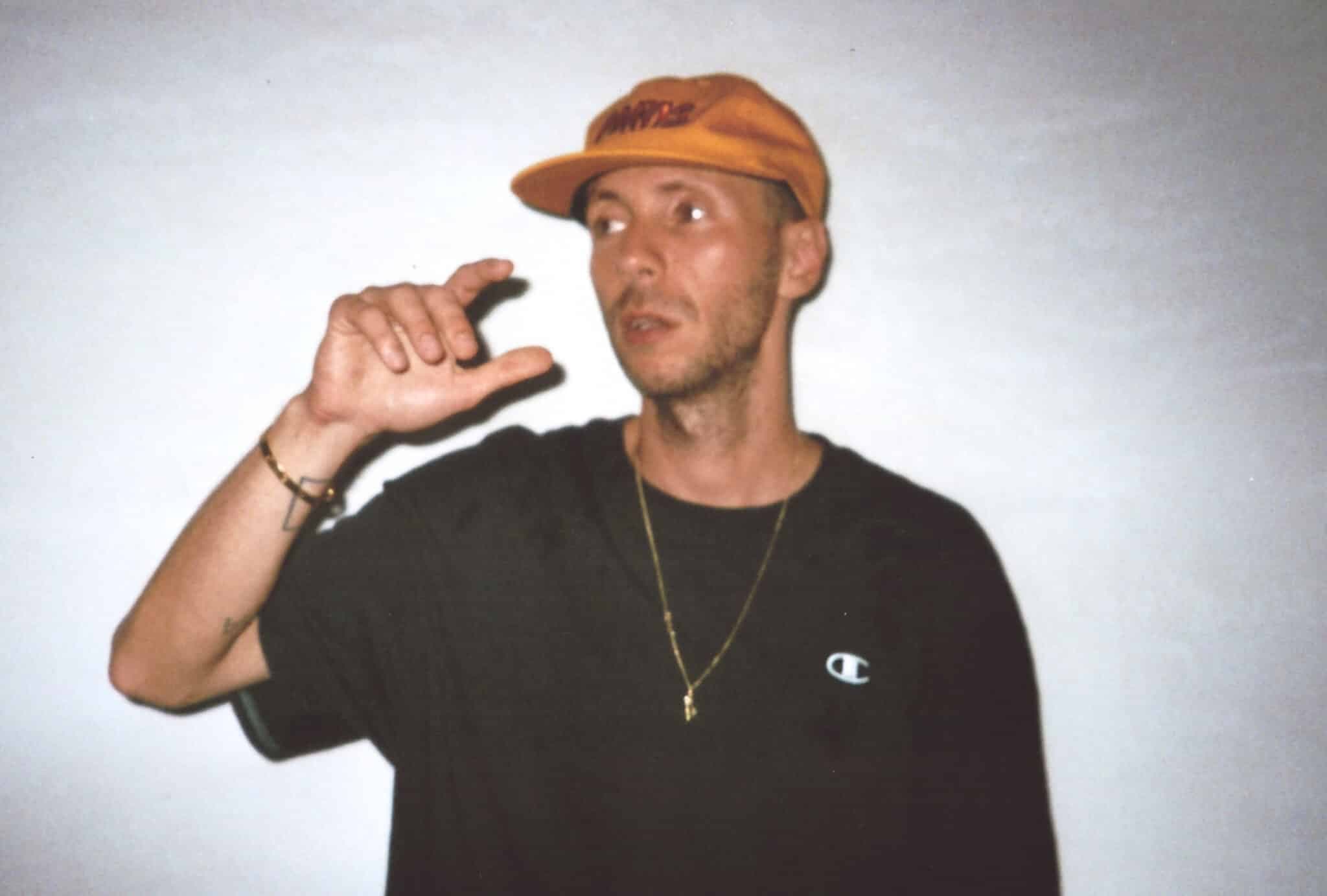 Man wearing an orange cap and black shirt gestures with one hand while looking to the side
