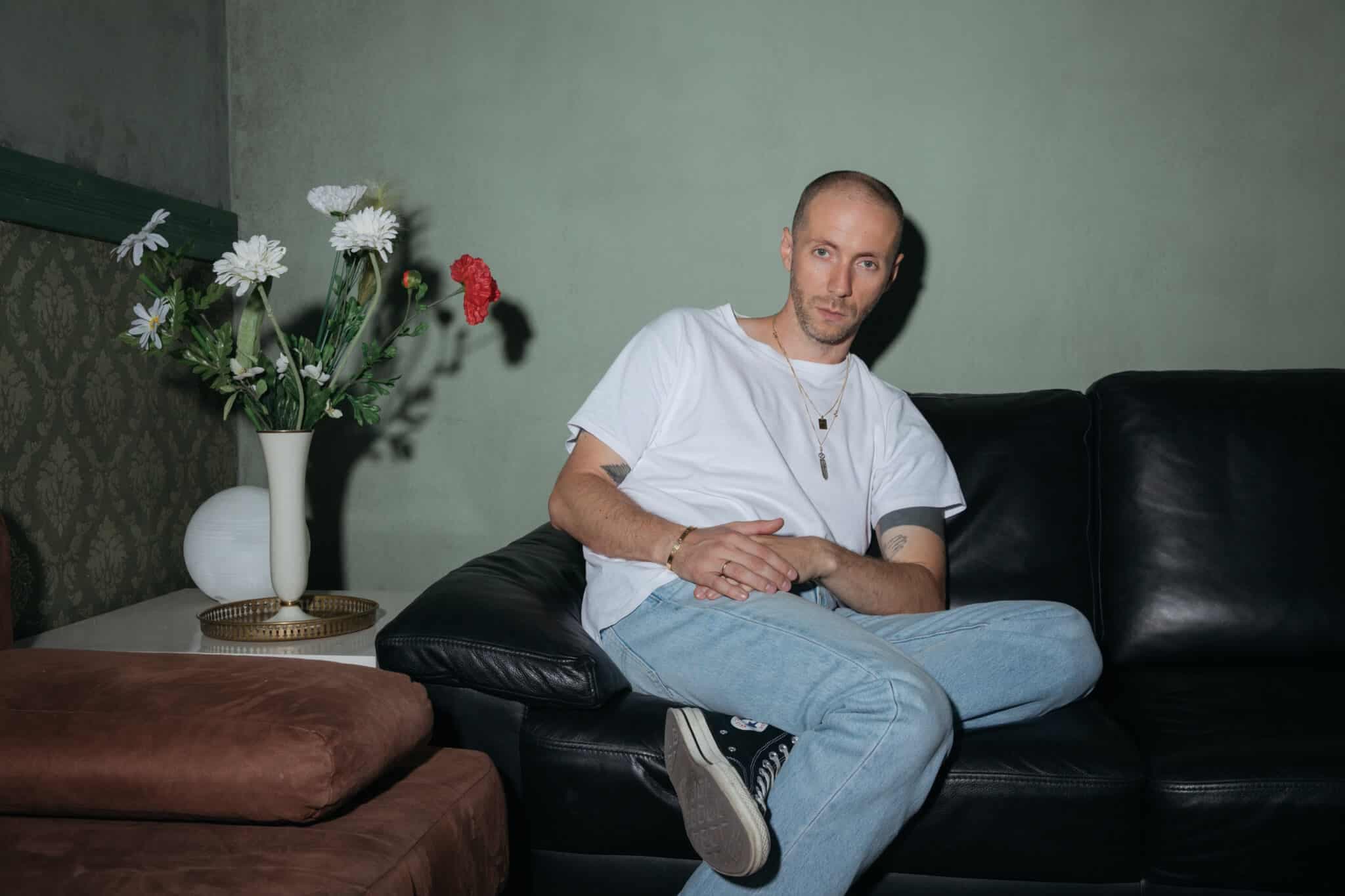 A man in a white T-shirt sits cross-legged on a black leather sofa beside a vase of flowers