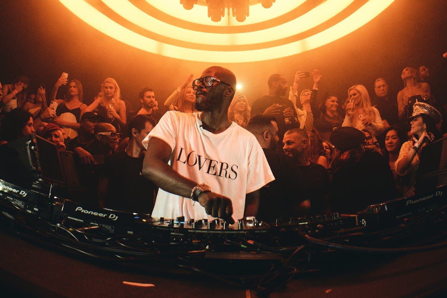 Black Coffee mixing music at a club booth as a crowd dances under orange lights
