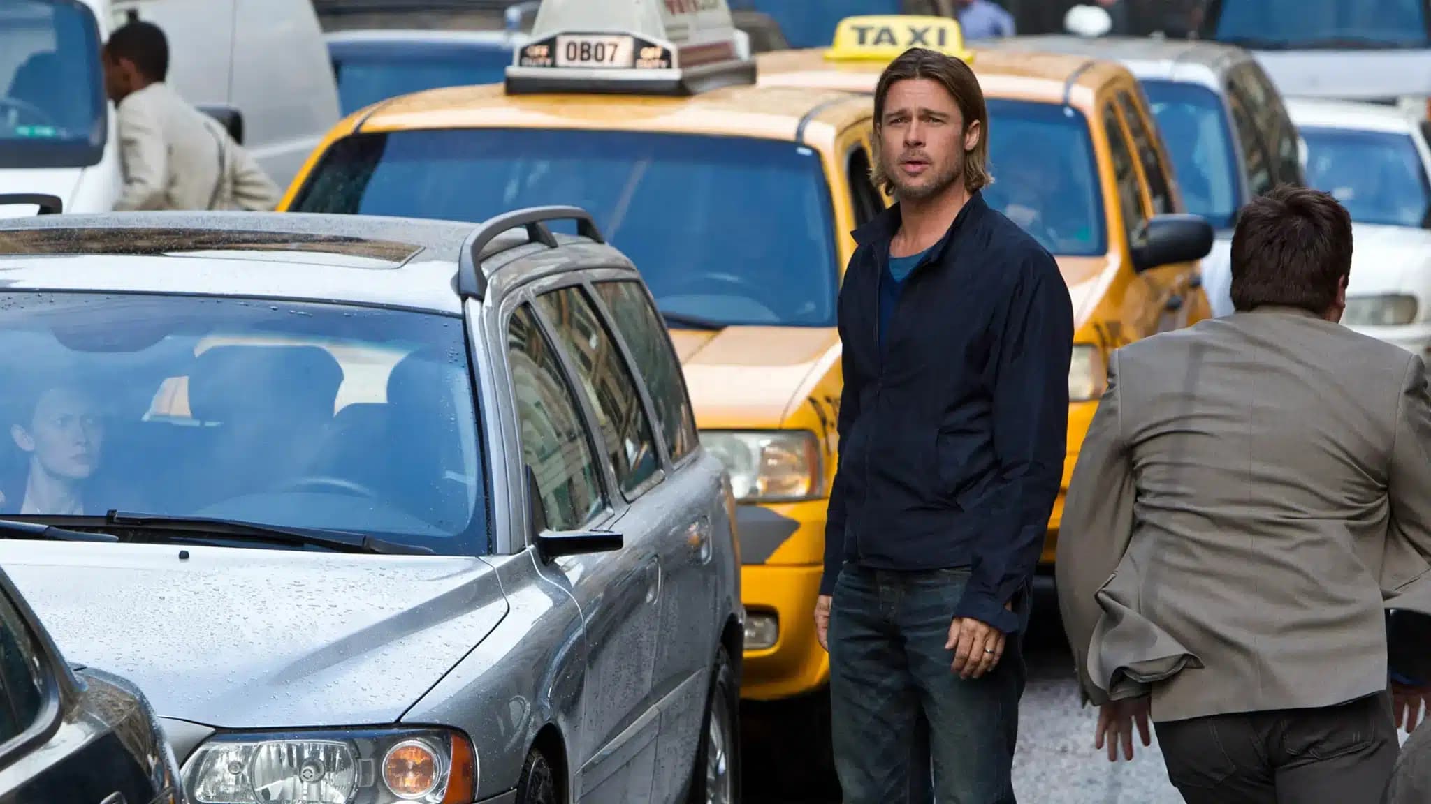 Brad Pitt stands beside a silver car amid yellow taxis, while another man in a suit faces him