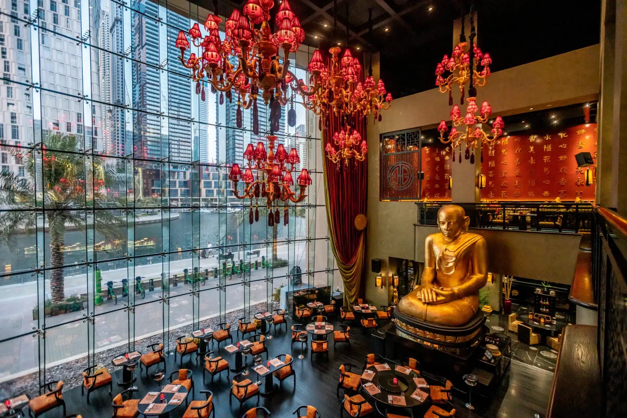 Upscale restaurant interior with red chandeliers, a large golden seated Buddha, and floor-to-ceiling windows overlooking city skyscrapers