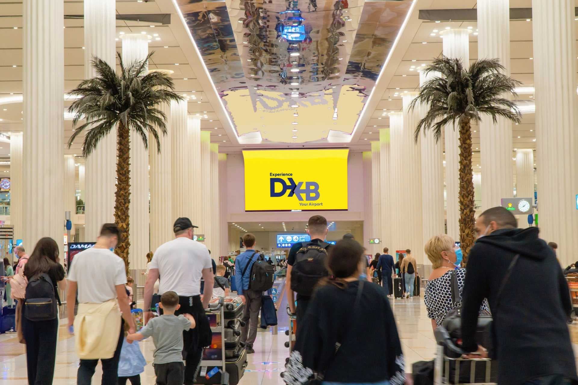 Travelers with luggage walk through Dubai International Airport beneath palm trees and a sign reading “Experience DXB Your Airport.”