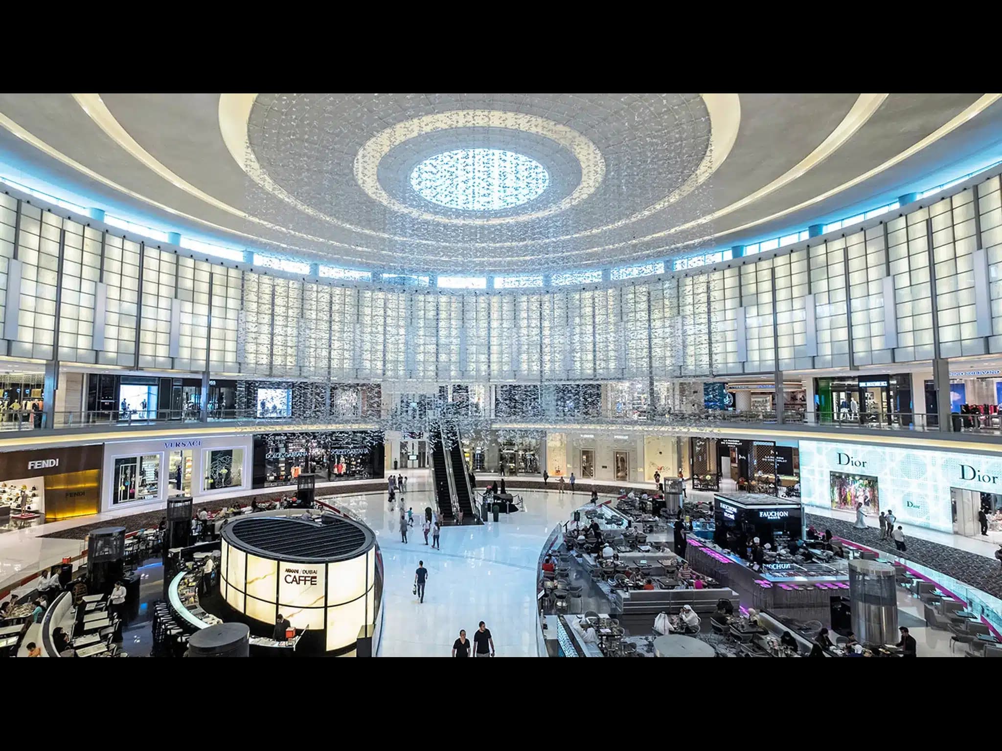 Grand luxury shopping mall atrium with circular skylight, escalators, cafés, and designer stores including Fendi, Versace, and Dior