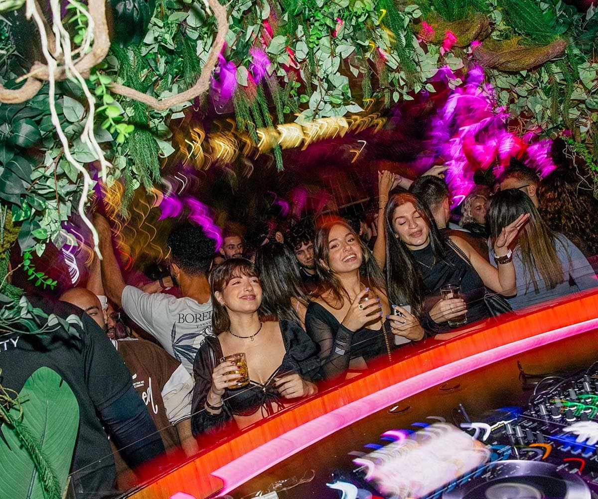Crowd dancing beneath lush greenery and vivid purple lights at a nightclub, with a DJ booth in the foreground