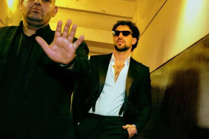 Two men in a dim hallway; the foreground man raises his hand toward the camera while the other wears sunglasses and a suit
