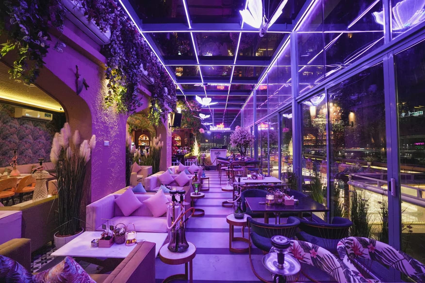 Purple-lit restaurant lounge with plush seating, tables, hookahs, hanging plants, and floor-to-ceiling windows overlooking the city