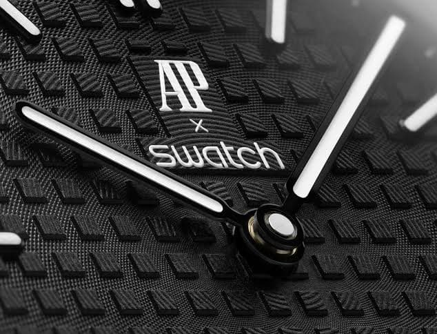 Close-up of a black textured watch dial with white hands and “AP x Swatch” branding