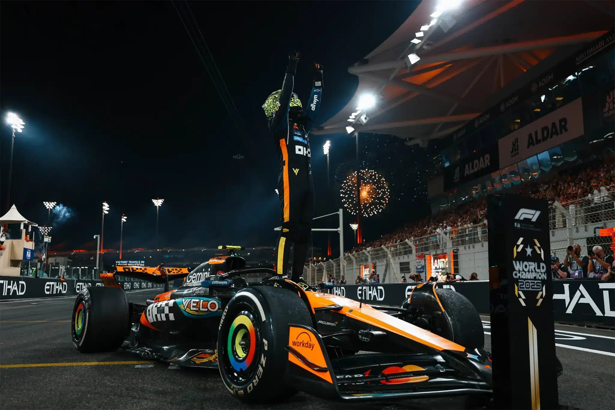 McLaren Formula 1 driver celebrates atop the car after a night race, with fireworks and cheering spectators in the background