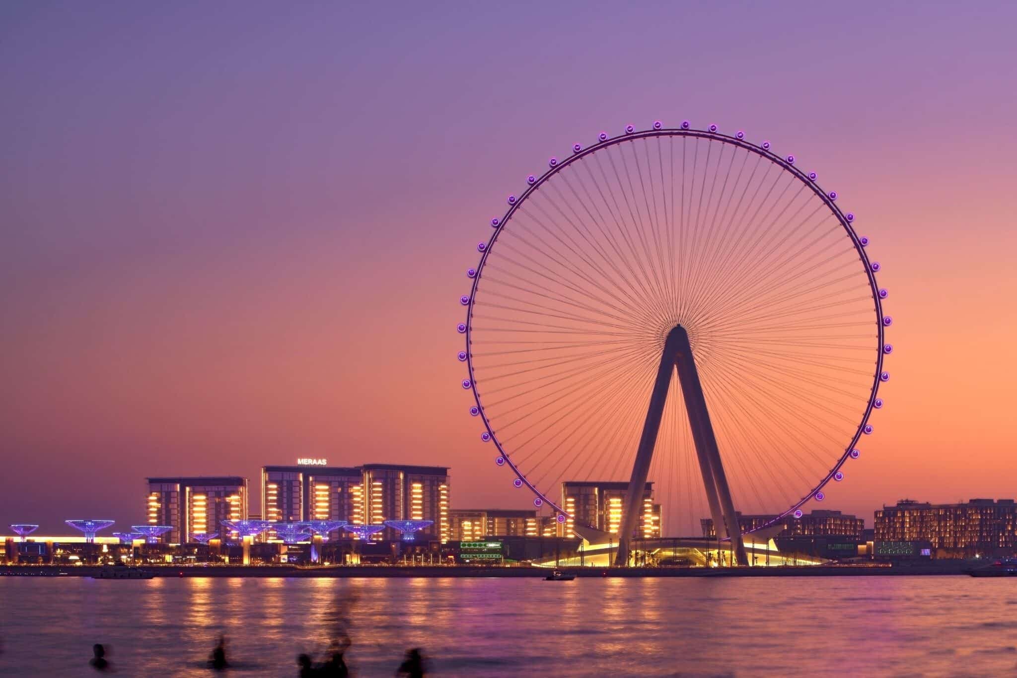A giant illuminated Ferris wheel and waterfront buildings at dusk, reflected in the water