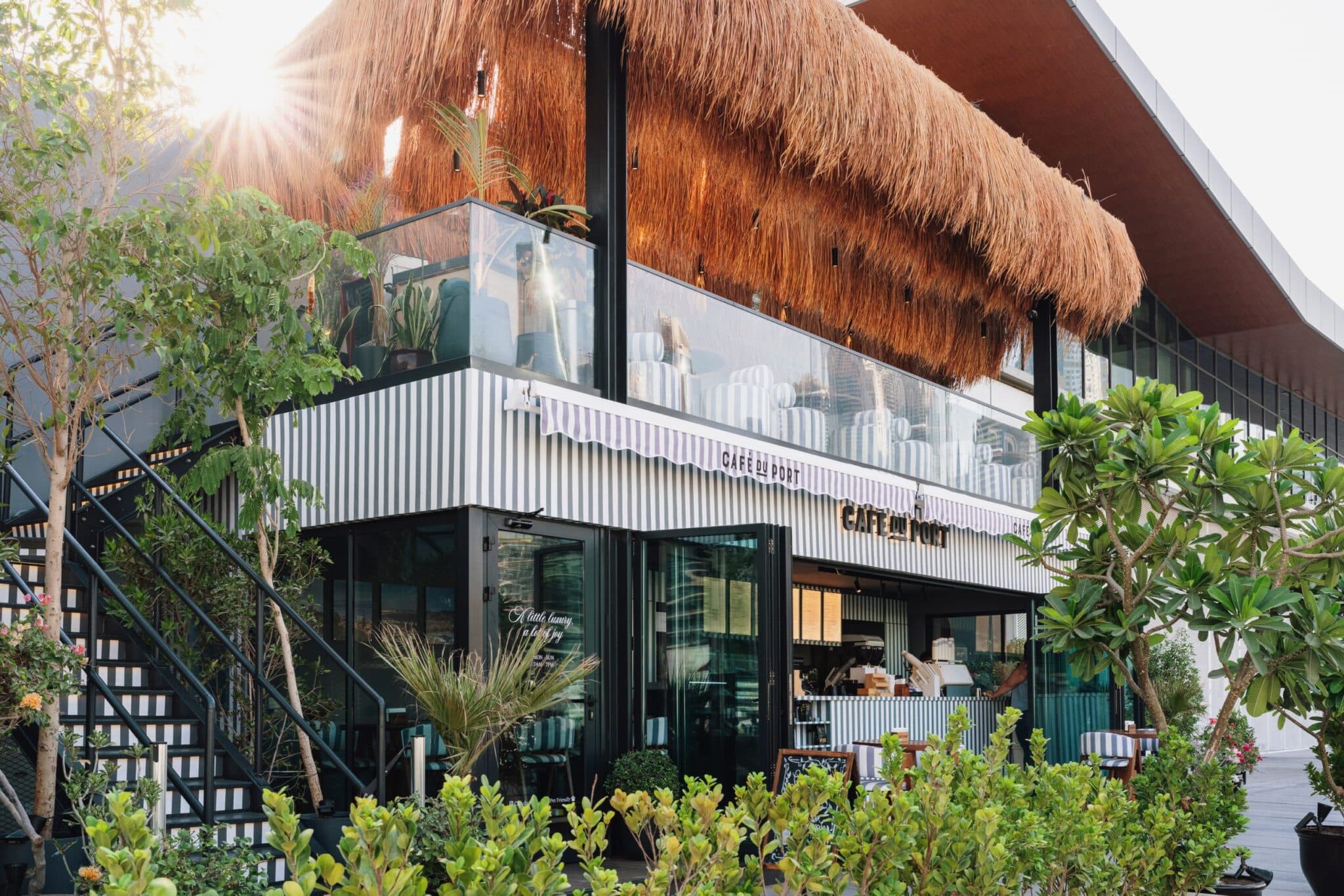 Café du Port with striped awnings, glass walls, tropical plants, and a thatched-roof terrace