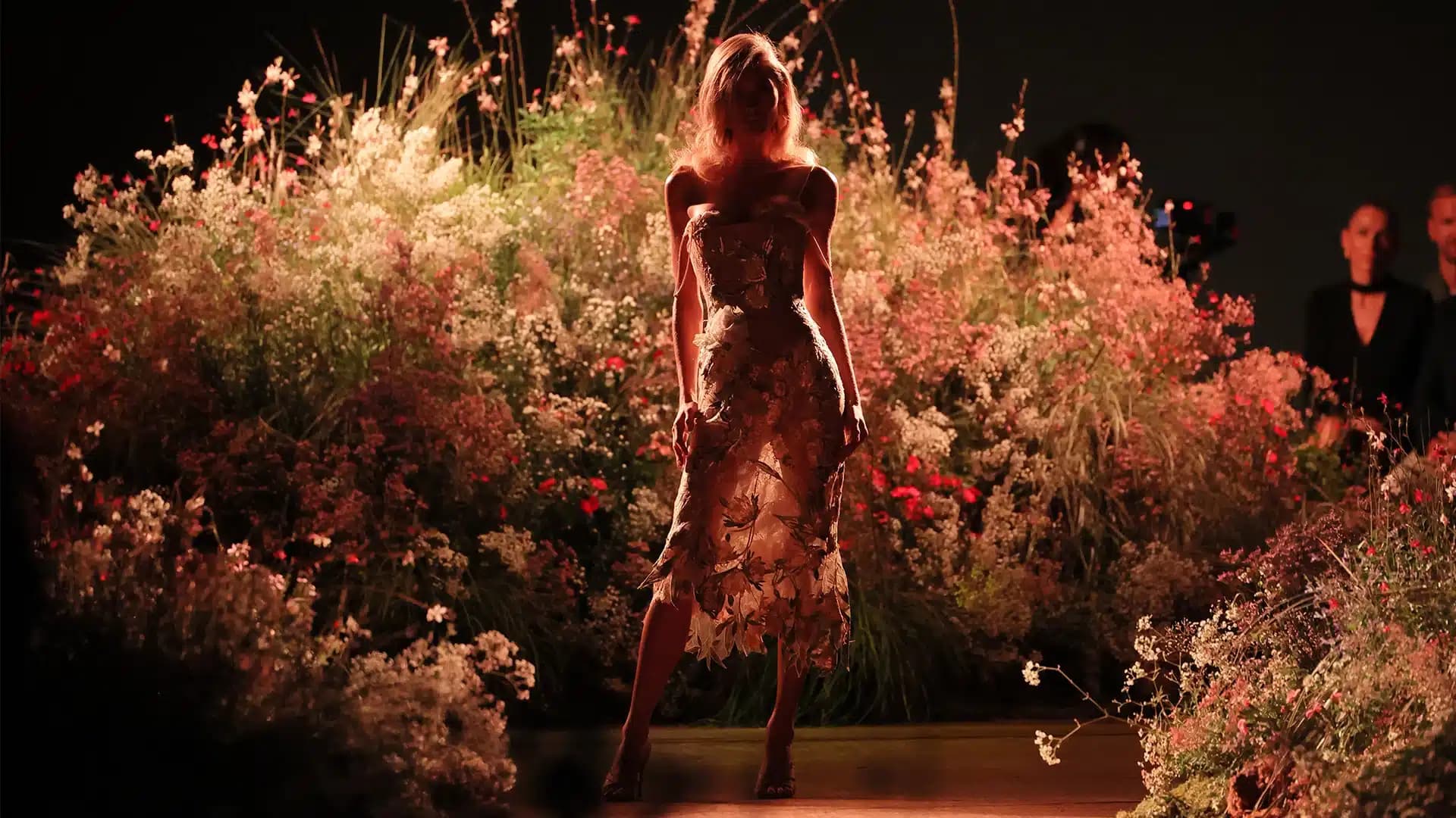 Model in a floral-patterned dress standing on a runway surrounded by illuminated flowers