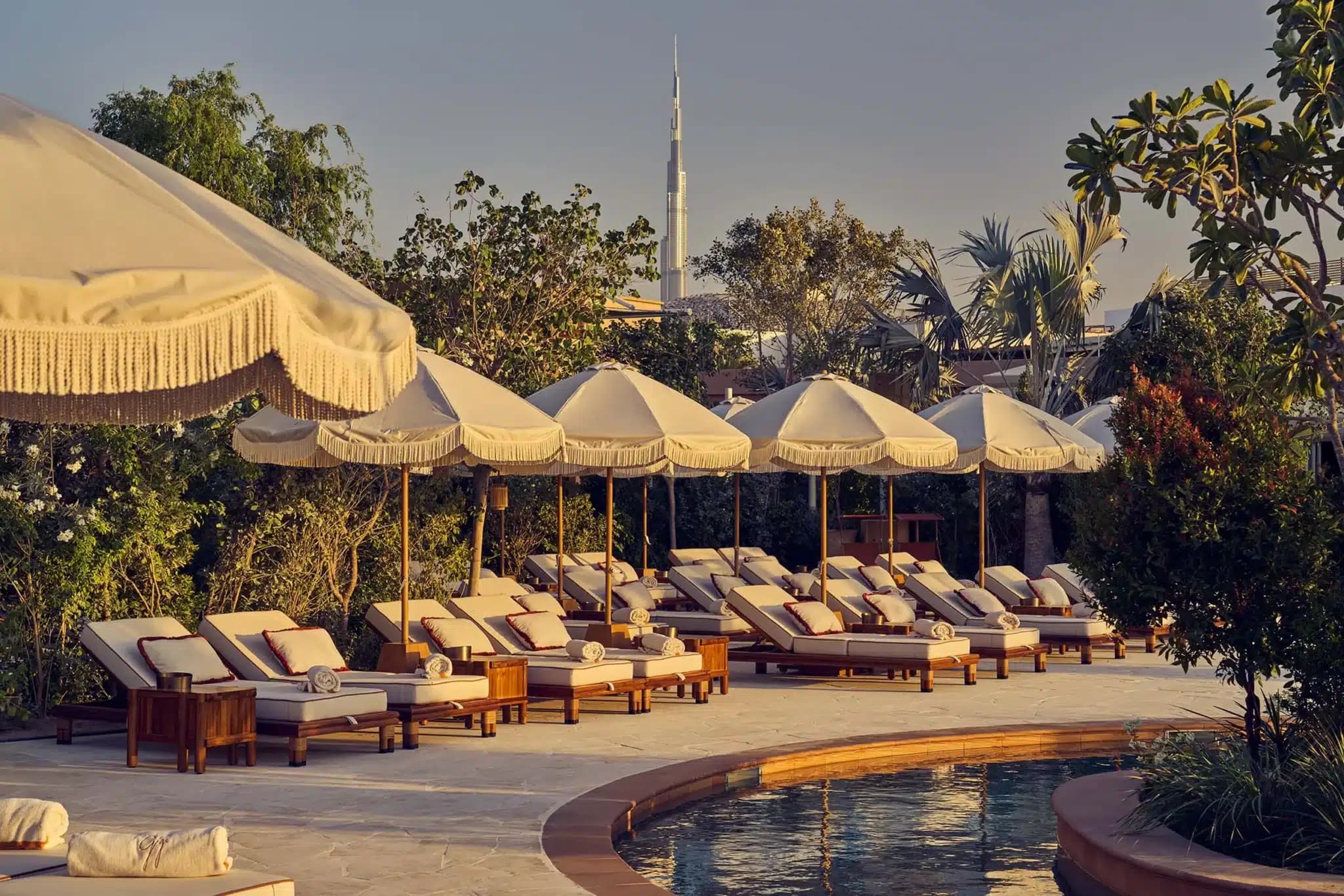 Poolside lounge with cream fringed umbrellas and cushioned loungers, with Dubai’s Burj Khalifa visible beyond the gardens