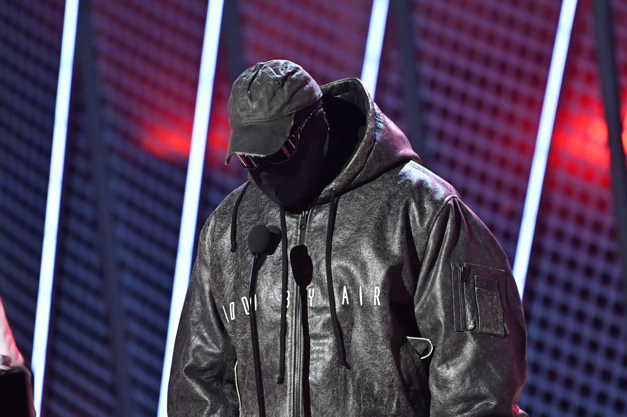 Masked performer wearing a cap and black hoodie on a brightly lit stage