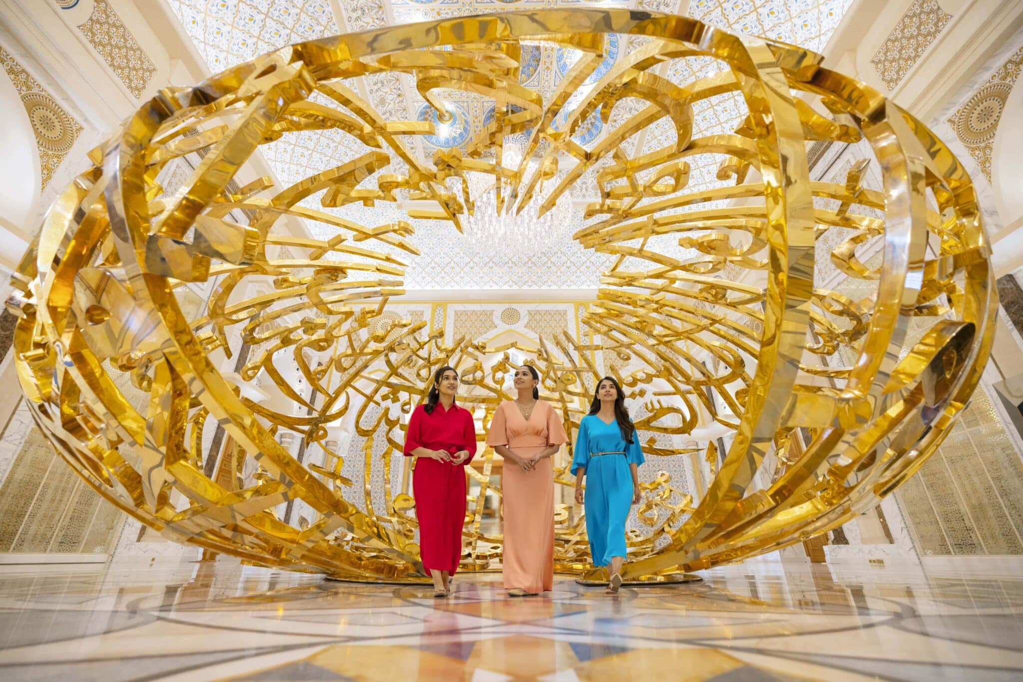 Three women in colorful dresses stand beneath a large golden calligraphic sculpture in an ornate, patterned hall
