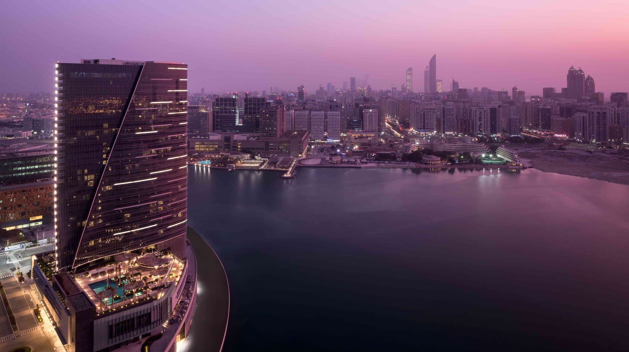 Twilight aerial view of Abu Dhabi’s waterfront skyline, with illuminated high-rises and a modern hotel complex in the foreground