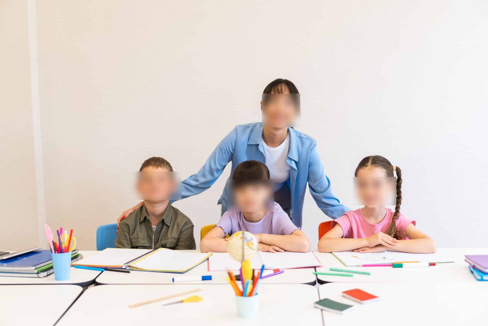 Teacher standing behind three children seated at desks with notebooks, pencils, and a globe in a classroom