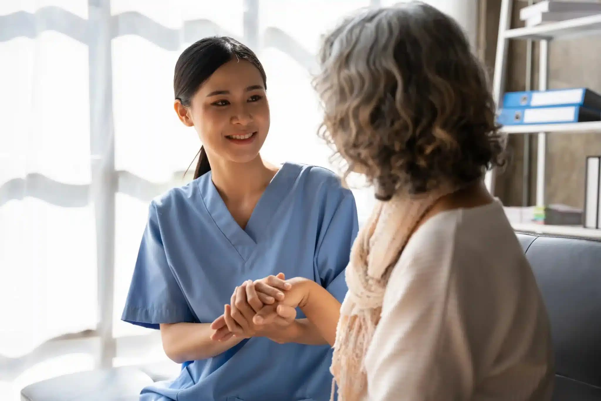A healthcare worker comforts an older woman by holding her hands in a bright care setting