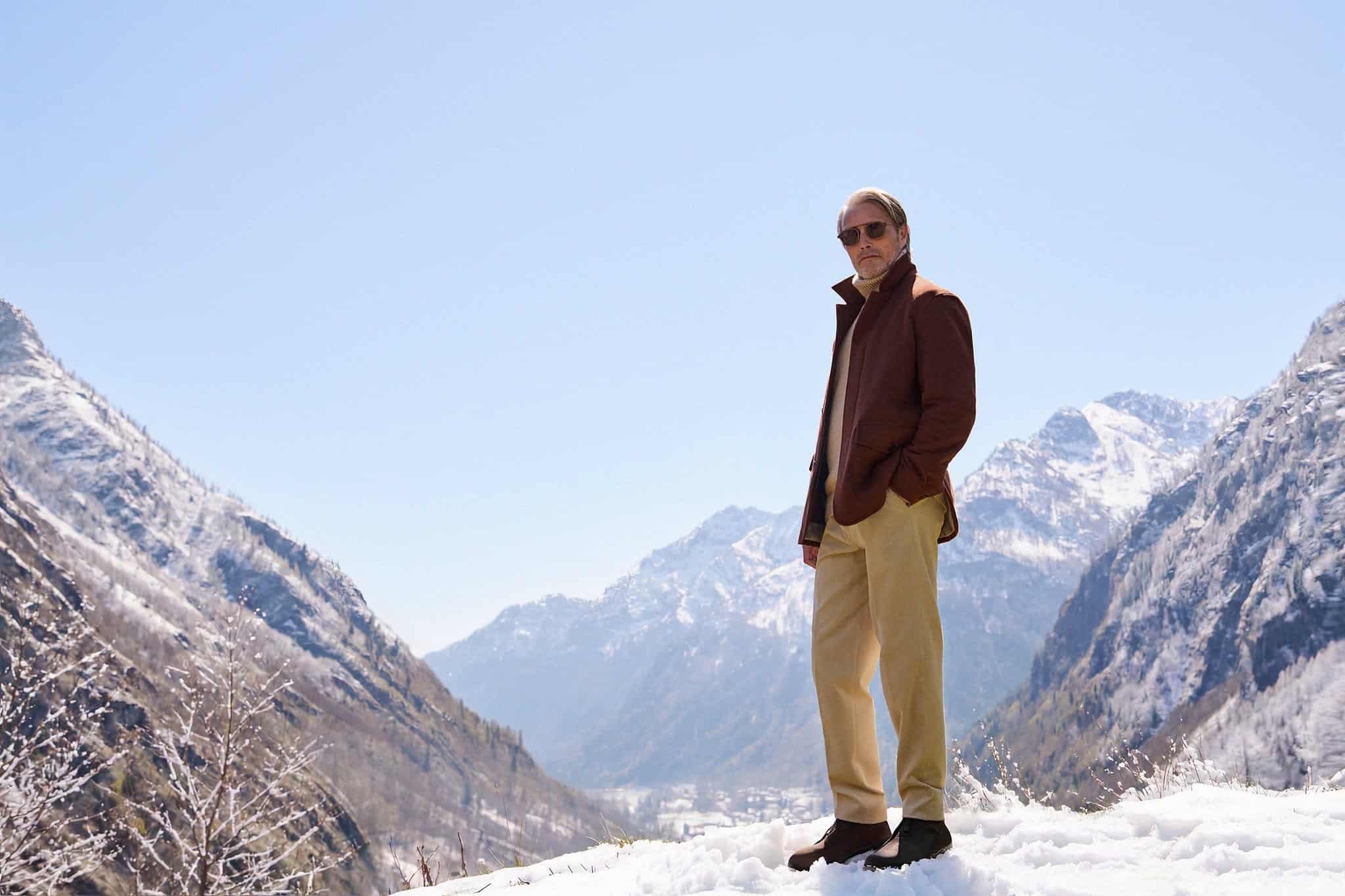 Man wearing sunglasses and a brown coat stands in a snowy mountain valley