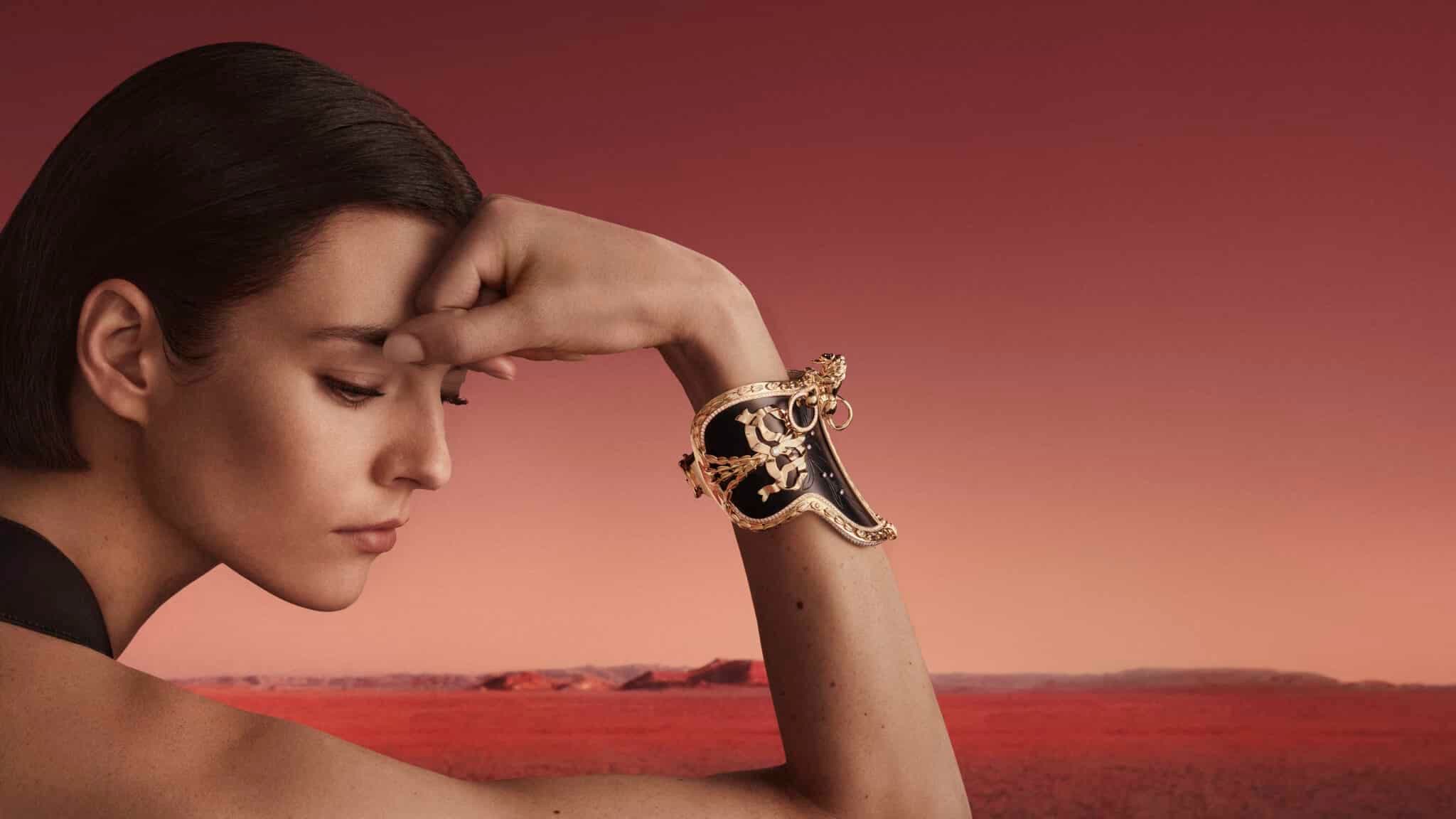 Woman wearing an ornate gold-and-black bracelet, resting her hand against her forehead against a red desert landscape