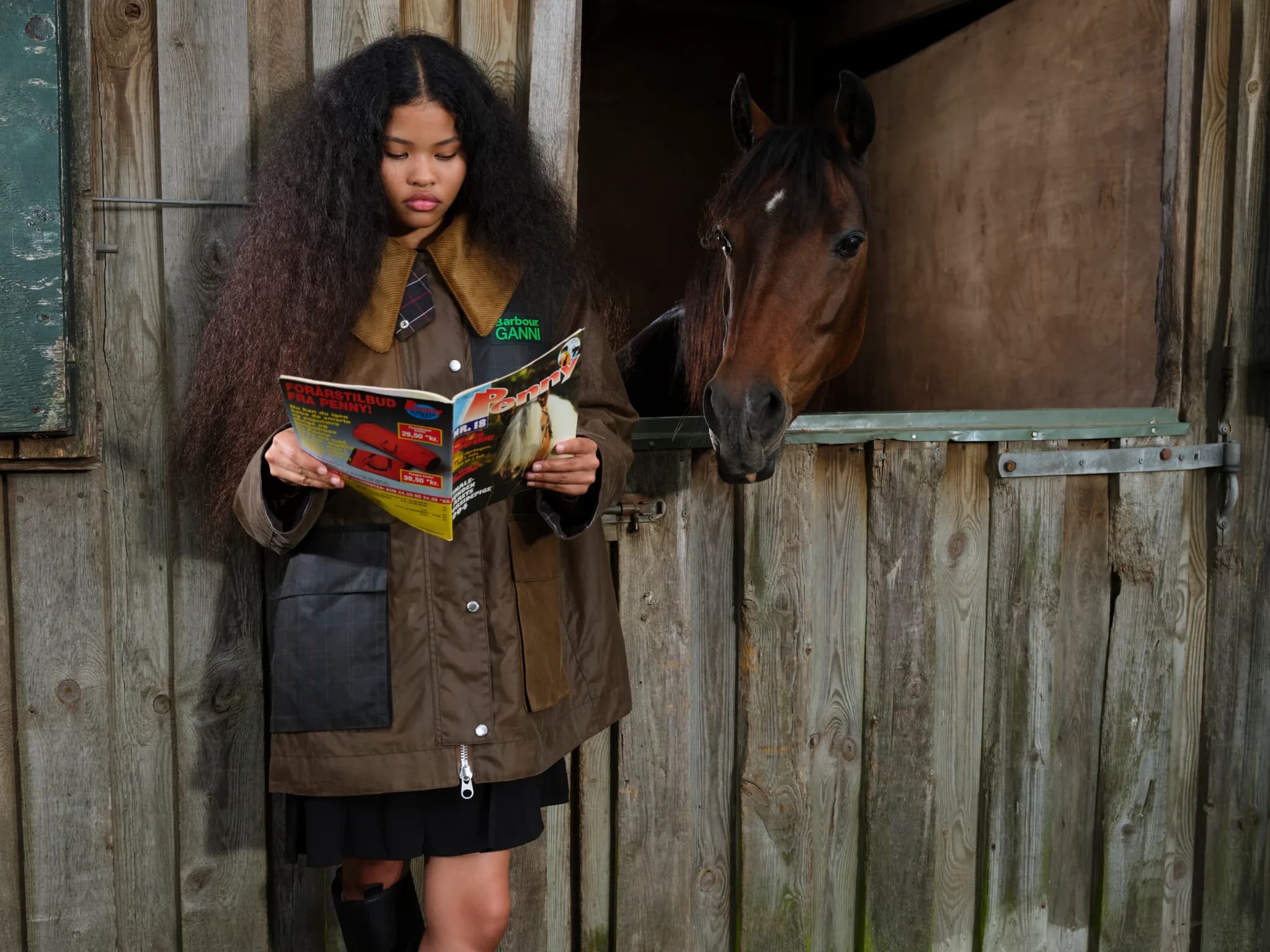 A woman in a brown riding jacket reads a magazine beside a horse leaning out of a wooden stable stall