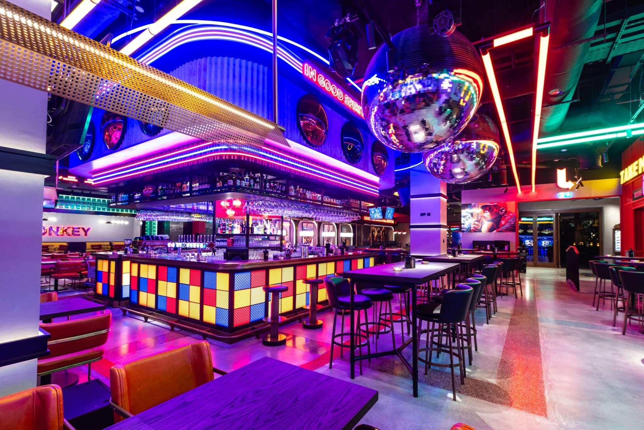 Neon-lit bar and restaurant interior with colorful seating, a central bar, disco balls, and vibrant geometric lighting