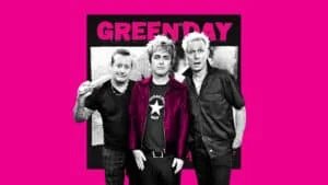 Three band members stand together in front of a pink background with the words "GREEN DAY" in bold letters. One person holds drumsticks, another wears a star-themed shirt and a red jacket, and the third sports a dark button-up shirt.