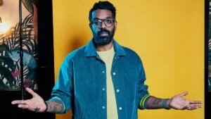 A man with glasses and a beard wearing a blue jacket and yellow shirt, standing against a bright yellow background with his arms outstretched.