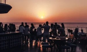 A group of people gathered on a balcony overlooking the ocean, silhouetted against a sunset in the background.