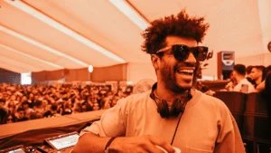 A smiling DJ wearing sunglasses and headphones stands in front of a mixing console with a large crowd in the background under a tent.