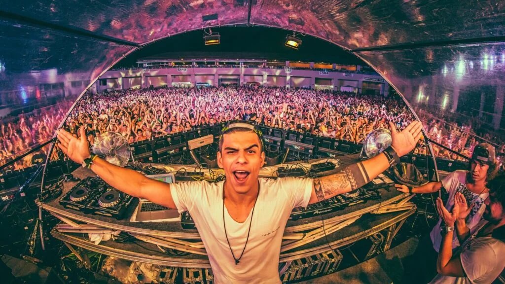 A DJ at a festival booth with arms wide open, smiling energetically towards a large, cheering crowd under a colorful, illuminated stage canopy.